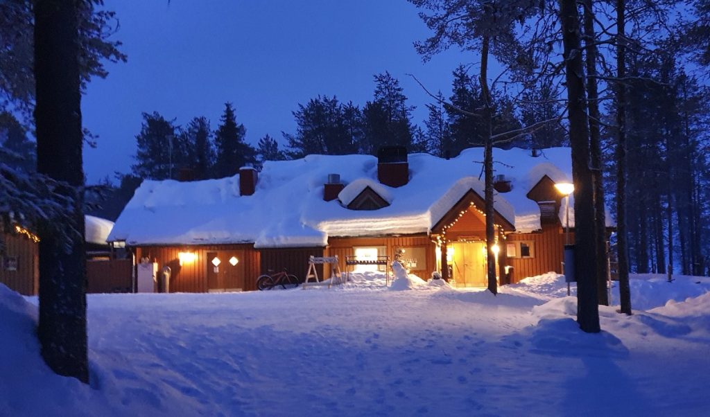 A warmly lit wooden cabin sits under deep snow in a pine forest at dusk, showing the cozy side of things to do in Finnish Lapland.