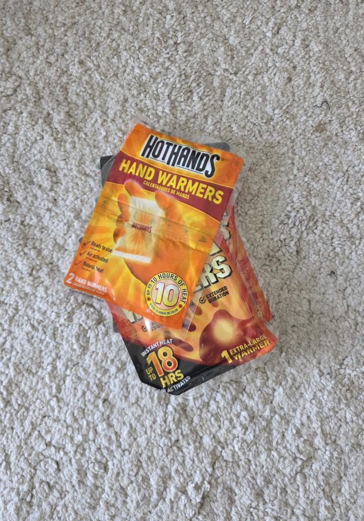 HotHands hand warmers and an extra large warmer packet laid on a cream carpet. The packaging includes the text “HotHands” “Hand Warmers” “2 hand warmers” “Up to 10 hours of heat” “Ready to use” “Air activated” “Natural heat” “Instant heat” “Up to 18 hrs” and “1 extra large warmer.”