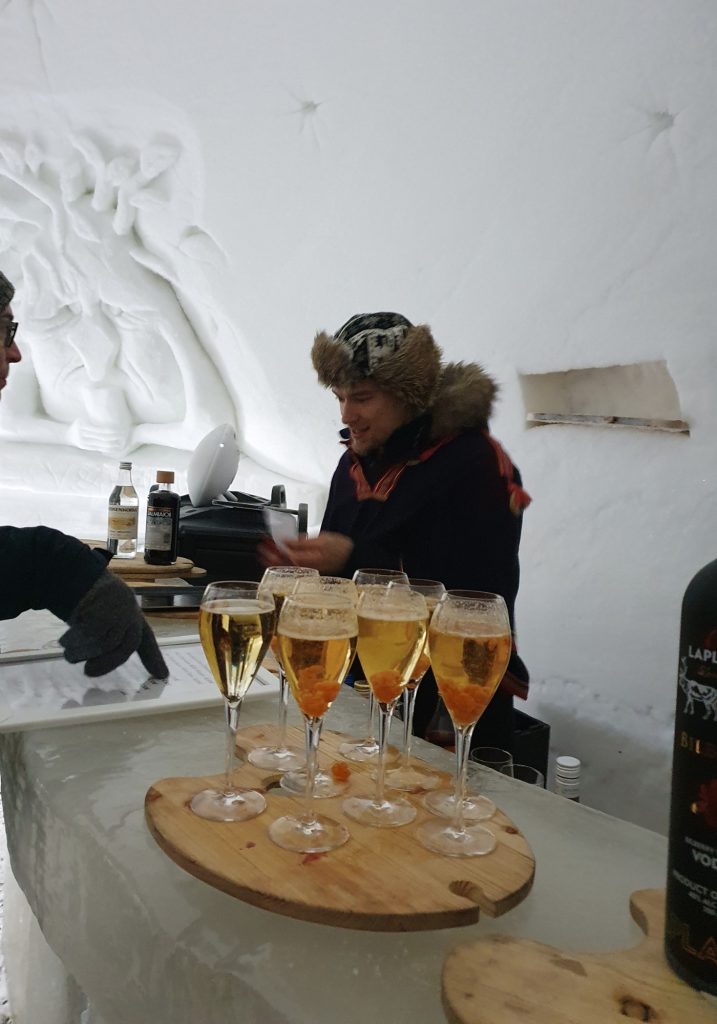 Several glasses of golden drinks sit on an ice bar inside a snow room, with a guide in a fur hat standing behind them.