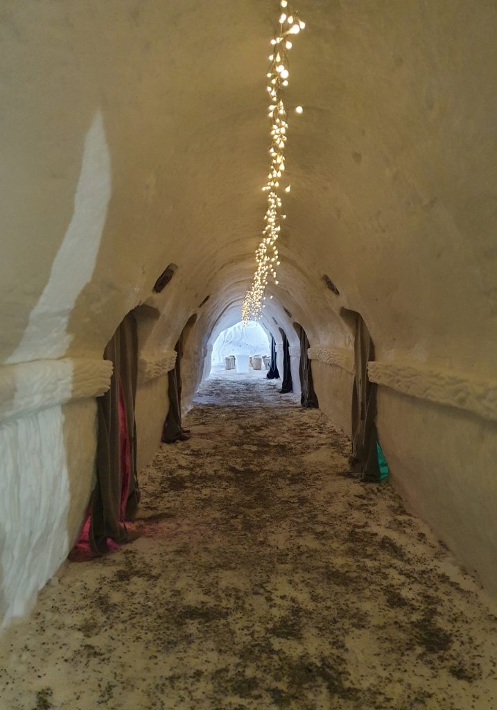 A snow tunnel hallway curves under warm string lights, with curtained alcoves built into the icy walls.