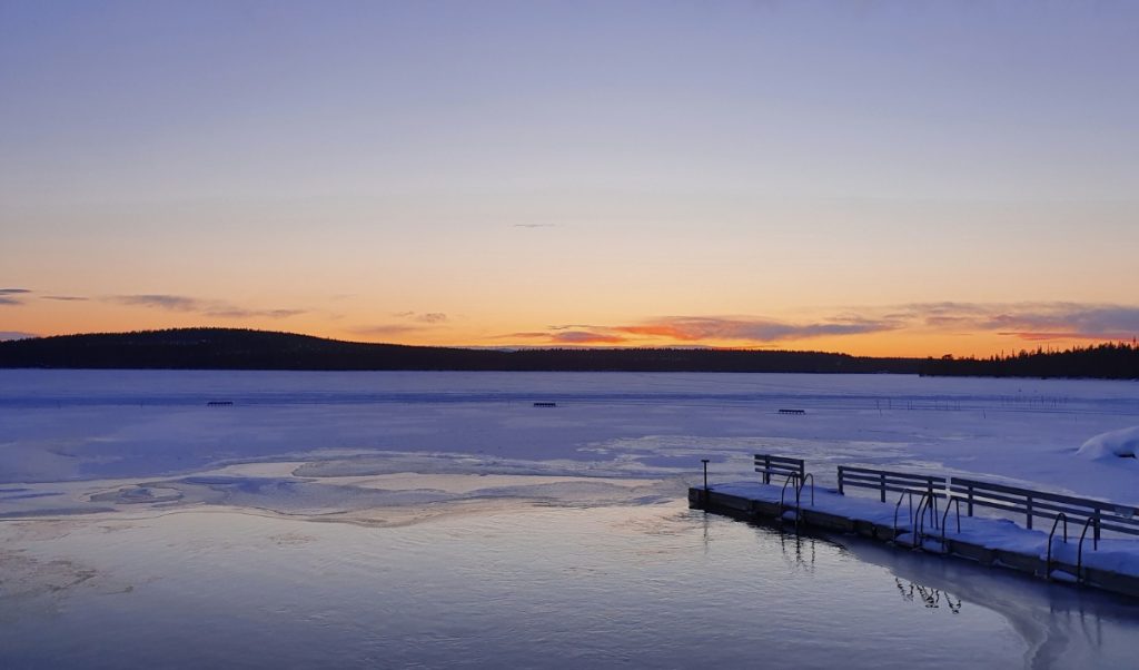 A wooden dock sits beside a partially frozen lake at sunset, with orange and purple sky over the distant forest.