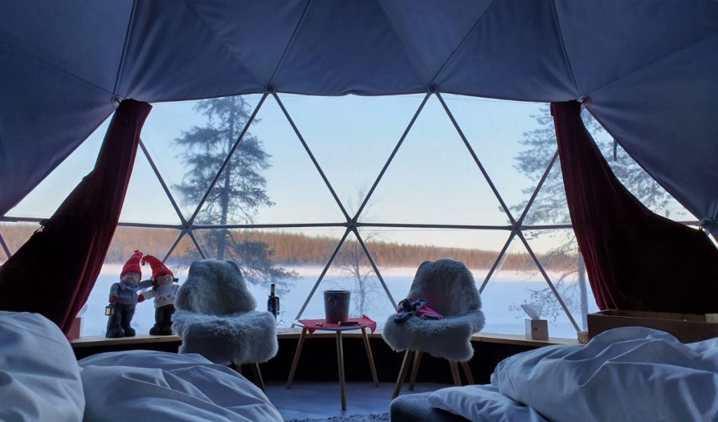A geodesic dome bedroom looks out over a snowy lake through large triangular windows, with fur covered chairs and red curtains framing the view.