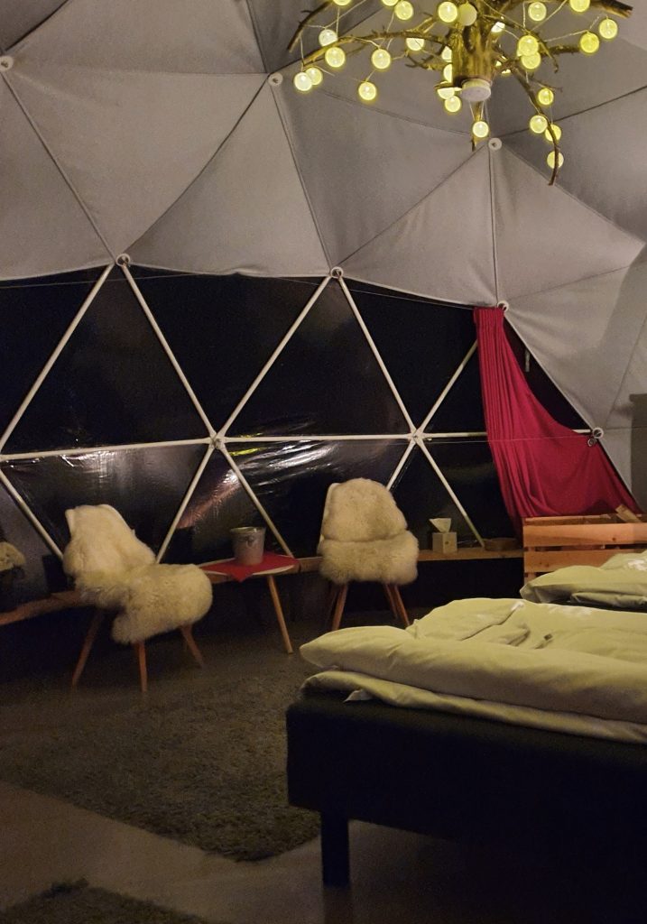 A cozy geodesic dome room has a bed, two fur covered chairs, red curtains, and a glowing branch chandelier overhead.