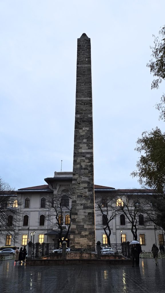 Tall stone Obelisk of Theodosius rising from a rainy square in Istanbul. Wet pavement reflects the lights from the surrounding buildings as people walk past with umbrellas.