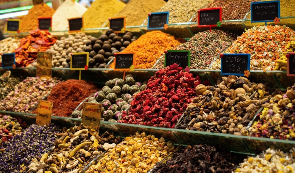 Colorful piles of spices and loose teas displayed in glass bins at an Istanbul market. Handwritten signs label items including "Jasmine Tea." "Mix Pepper." "Dried Vegetables." "Detox Tea." and "Energy Tea."