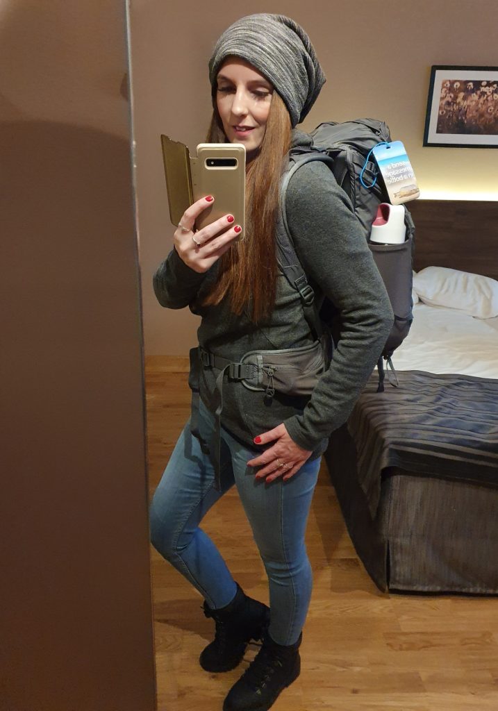 taking a mirror selfie in a hotel room while wearing a gray beanie, dark sweater, jeans, black boots, and a large travel backpack. A water bottle and luggage tag are tucked into the backpack, with an unmade bed behind her.