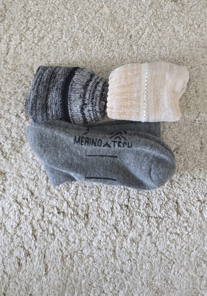 Four pairs of merino wool socks laid on a cream carpet. The set includes black and gray knit socks. Cream knit socks. And gray socks with “MERINOTECH” written across the top.