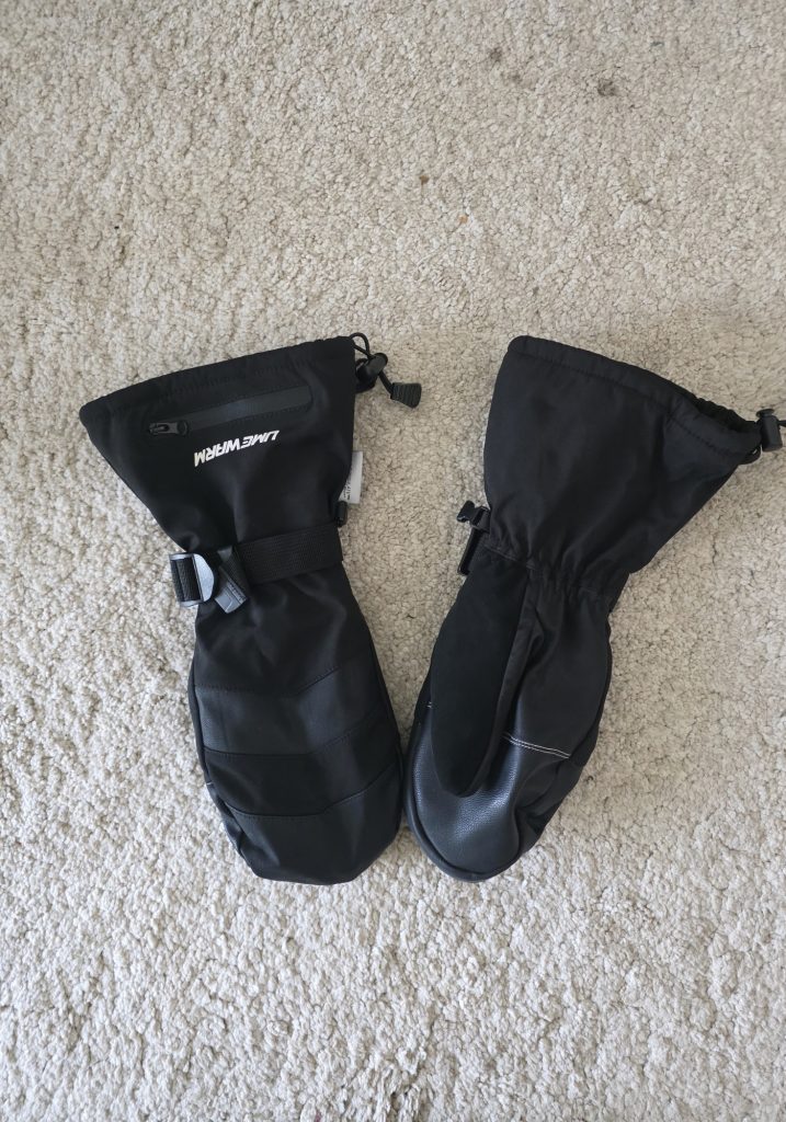 Black waterproof winter mittens laid on a cream carpet. One mitten has the brand name “LIMEWARM” and a small zipper pocket on the cuff.