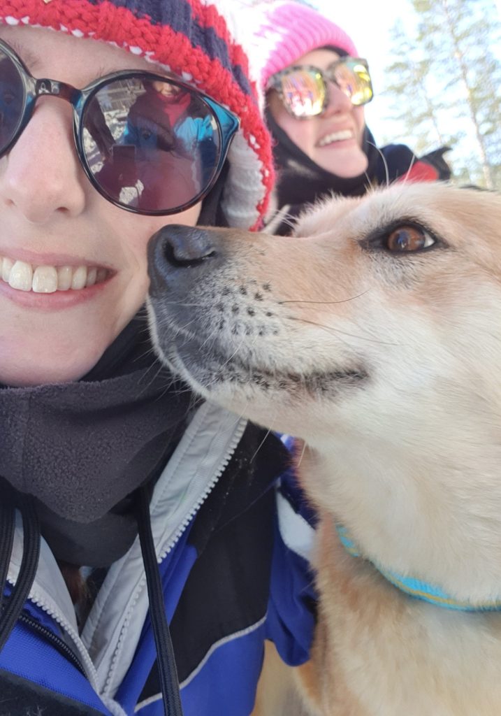 A sled dog presses its nose against a smiling woman bundled in winter gear, while another woman laughs in the snowy background.