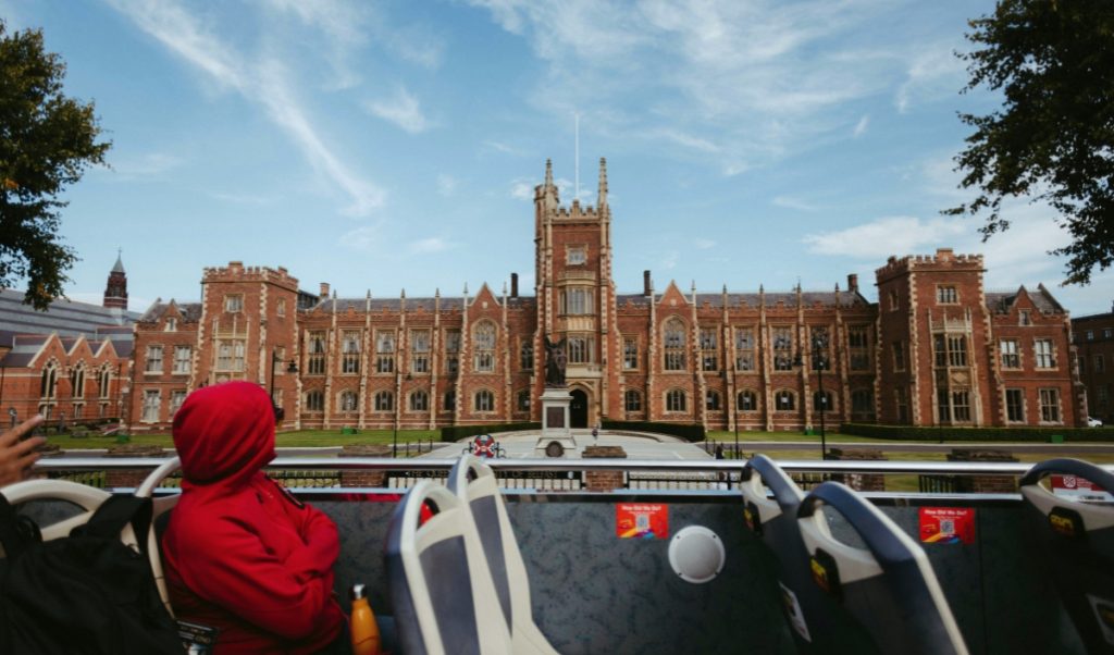 A person in a red hooded jacket sits on the top deck of a sightseeing bus facing a large red brick university building with a central tower. The scene shows a city tour passing historic architecture on a sunny day.