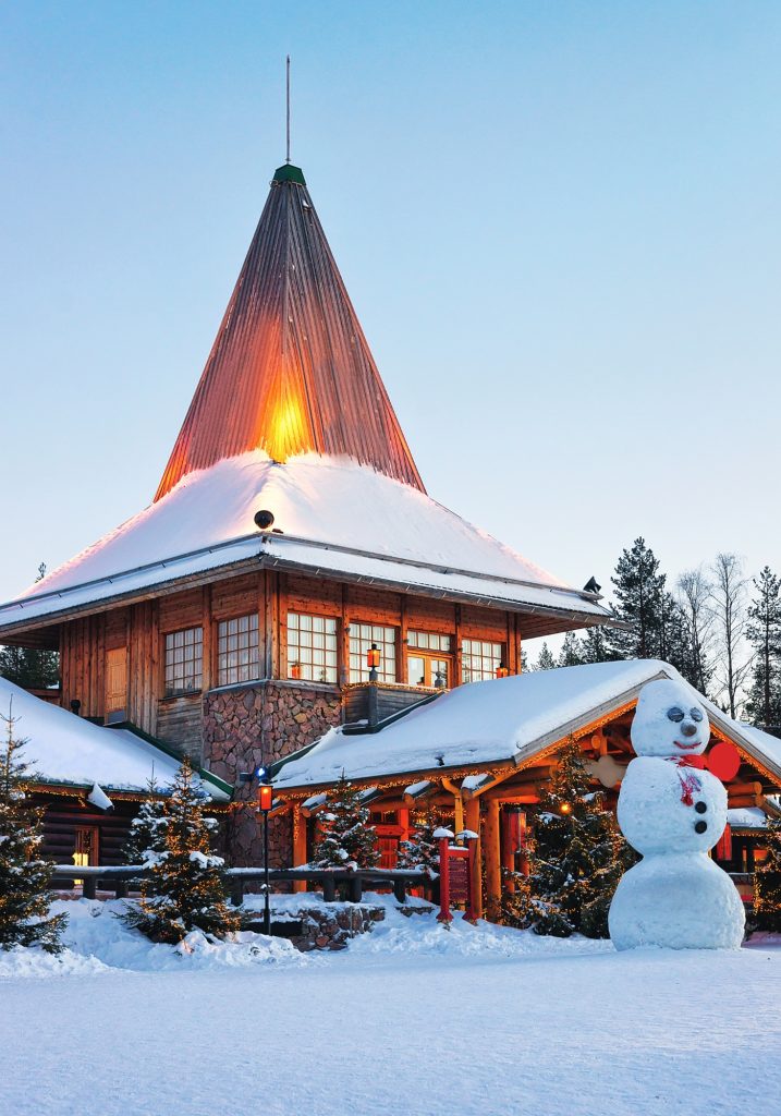 A tall wooden building with a steep glowing roof stands in the snow beside a large snowman and Christmas lights.