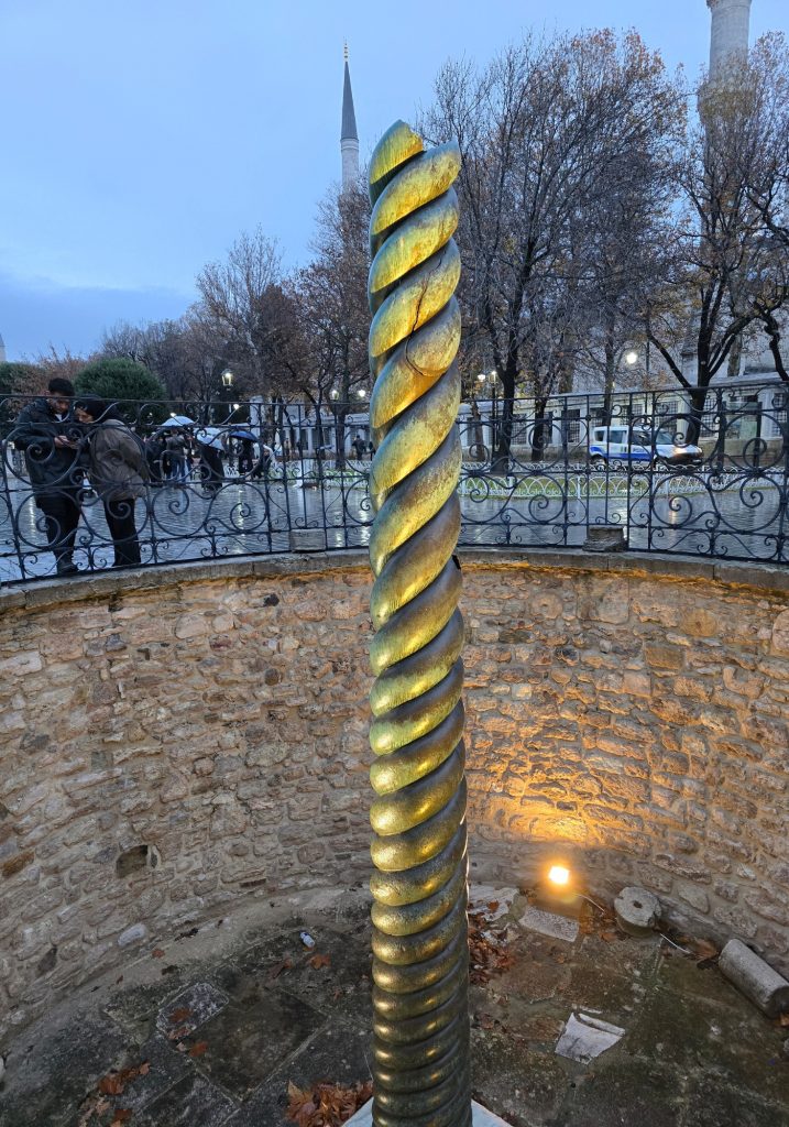 Serpent Column in Istanbul lit from below inside a stone-lined enclosure near the Hippodrome. A rainy street, bare trees, and mosque minarets appear behind the iron fence.