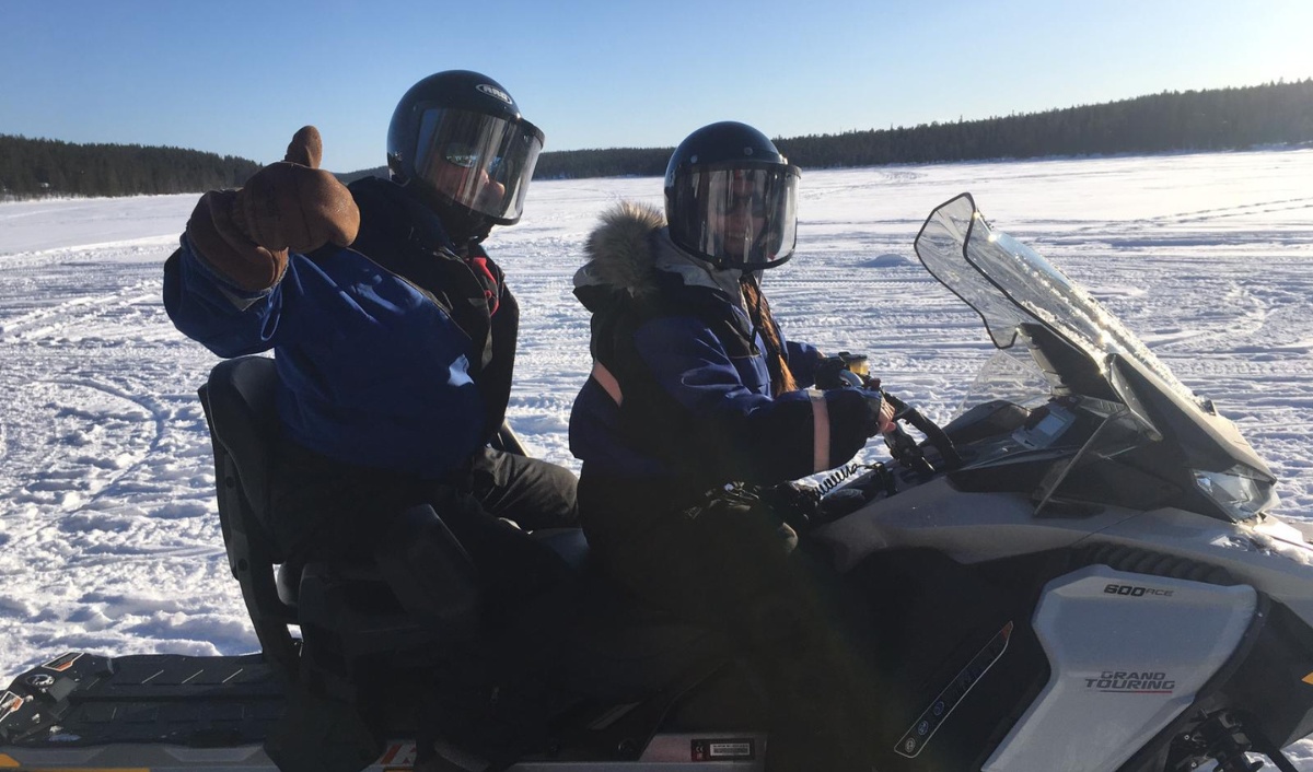 Two people in helmets and blue winter gear sit on a snowmobile on a frozen snowy lake, with one person giving a thumbs up.