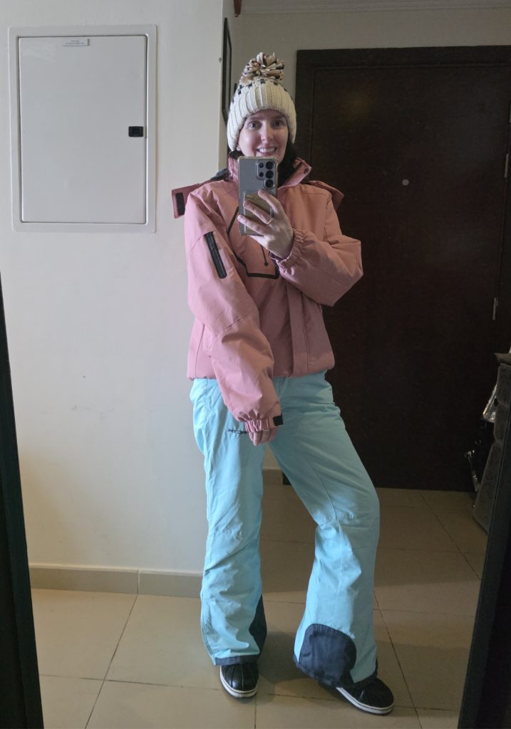 Taking a mirror selfie wearing a cream pom pom hat. Pink ski jacket. Light blue snow pants. And black winter snow boots.