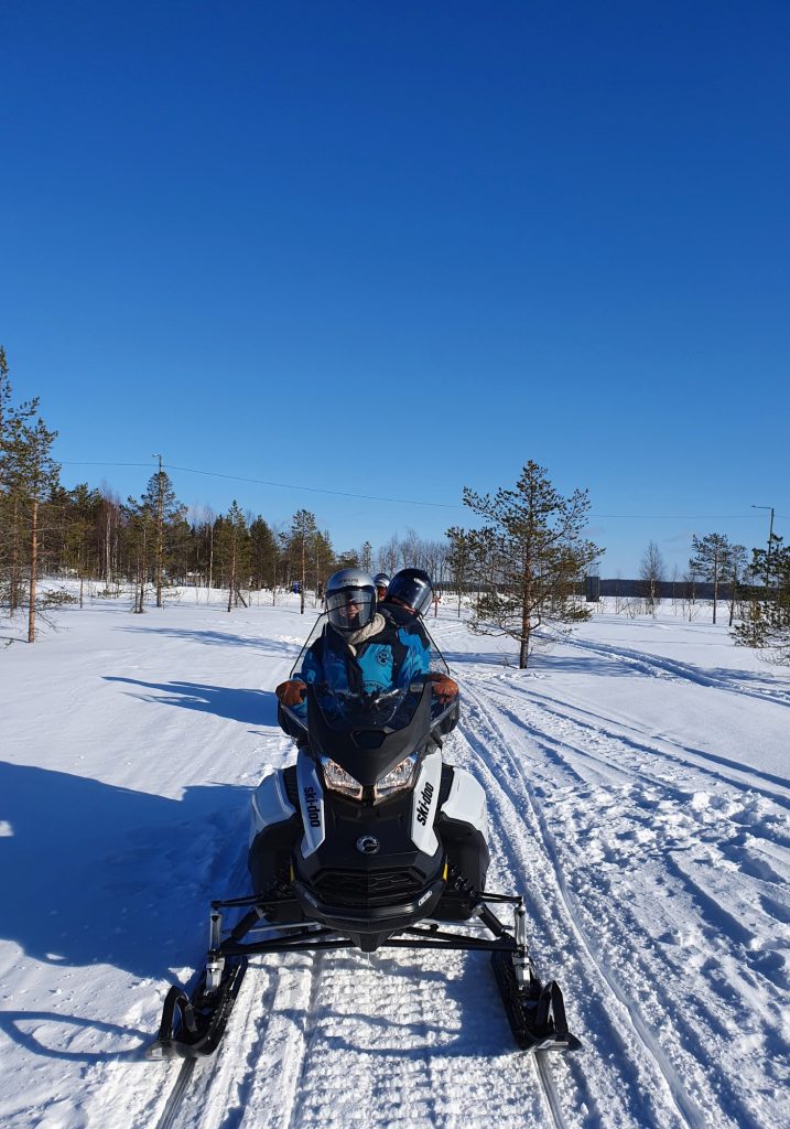 Two people in helmets ride a snowmobile along a snowy trail under a clear blue sky.
