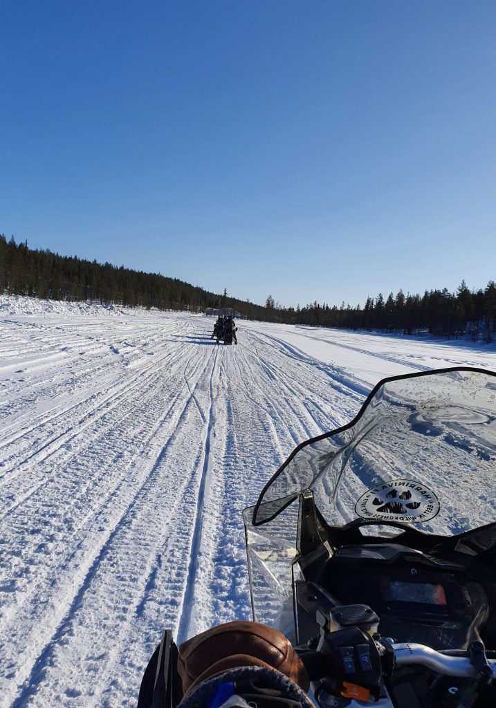 A snowmobile follows other riders across a wide frozen lake, with snow tracks stretching toward the forest ahead.
