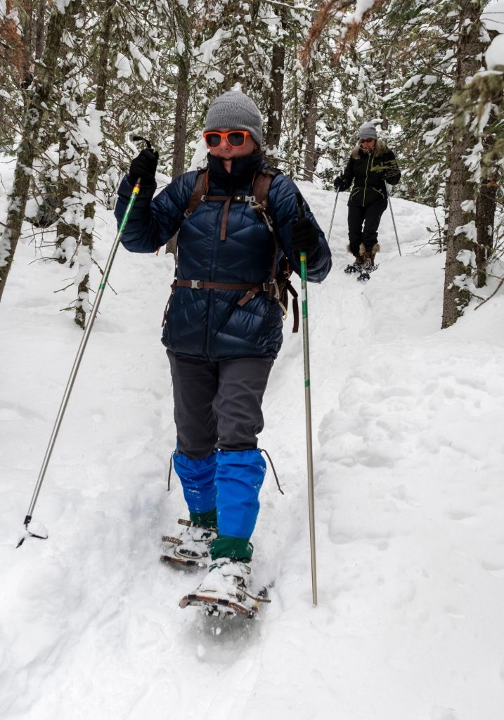 Two people snowshoe along a narrow trail through a snowy forest, using poles for balance.