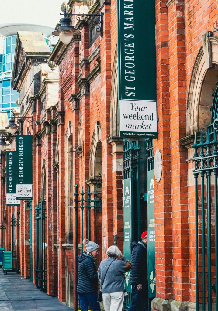 People stand outside a red brick market building with green signs reading "ST GEORGE'S MARKET" and "Your weekend market". The entrance and repeated hanging signs make the historic market feel like a key stop in the city.