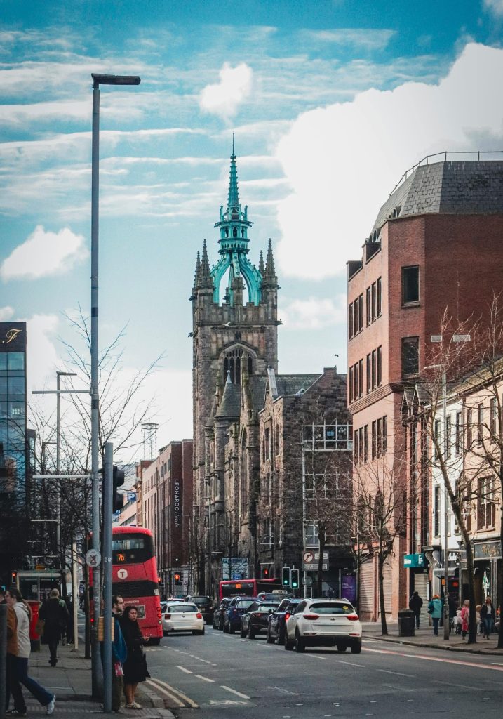 A busy city street with cars. pedestrians. and a red double decker bus leads toward an old stone church tower with a green spire.