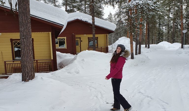 Me wearing a pink winter jacket and gray hat smiling while walking through deep snow beside red and yellow cabins in a snowy Finnish pine forest.