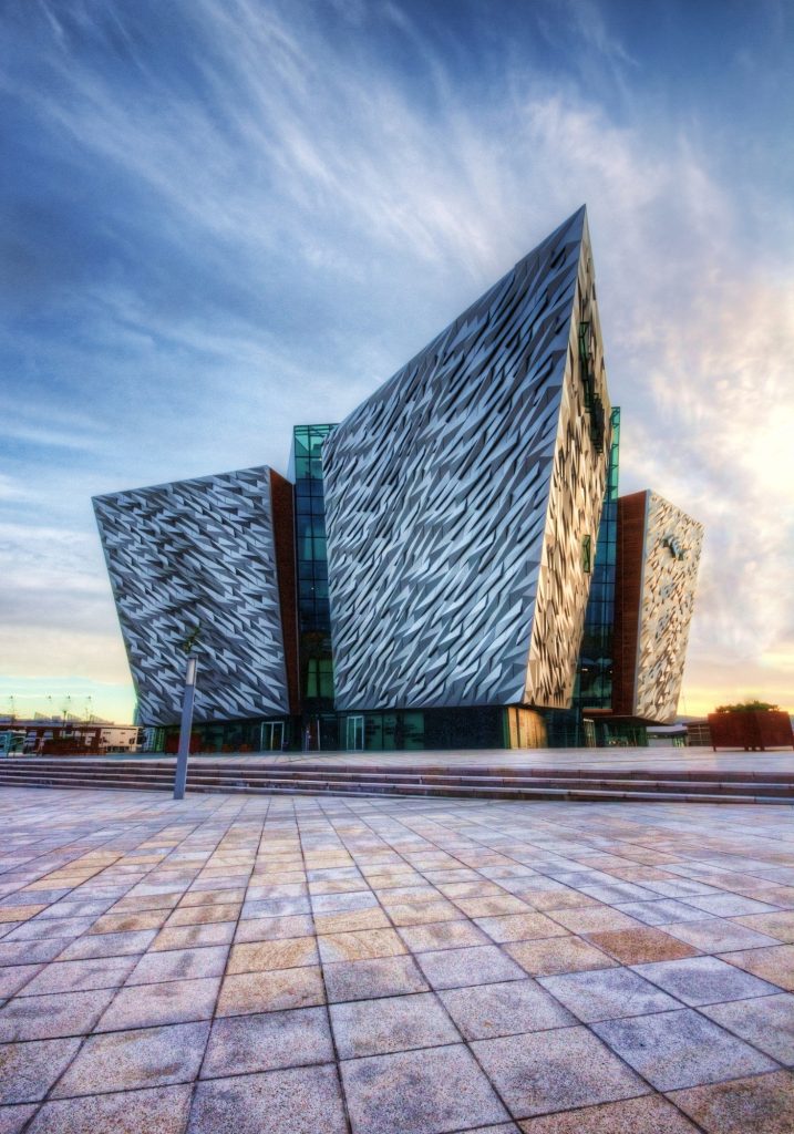 The Titanic Belfast building rises from a wide tiled plaza at sunset. Its sharp silver geometric panels resemble ship hulls and reflect the blue and gold sky.