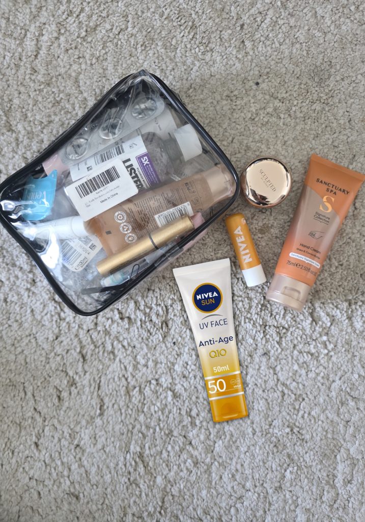 A clear toiletry bag filled with travel size products beside Nivea sunscreen. Nivea lip balm. A small round Sculp’d product. And Sanctuary Spa hand cream on a cream carpet.