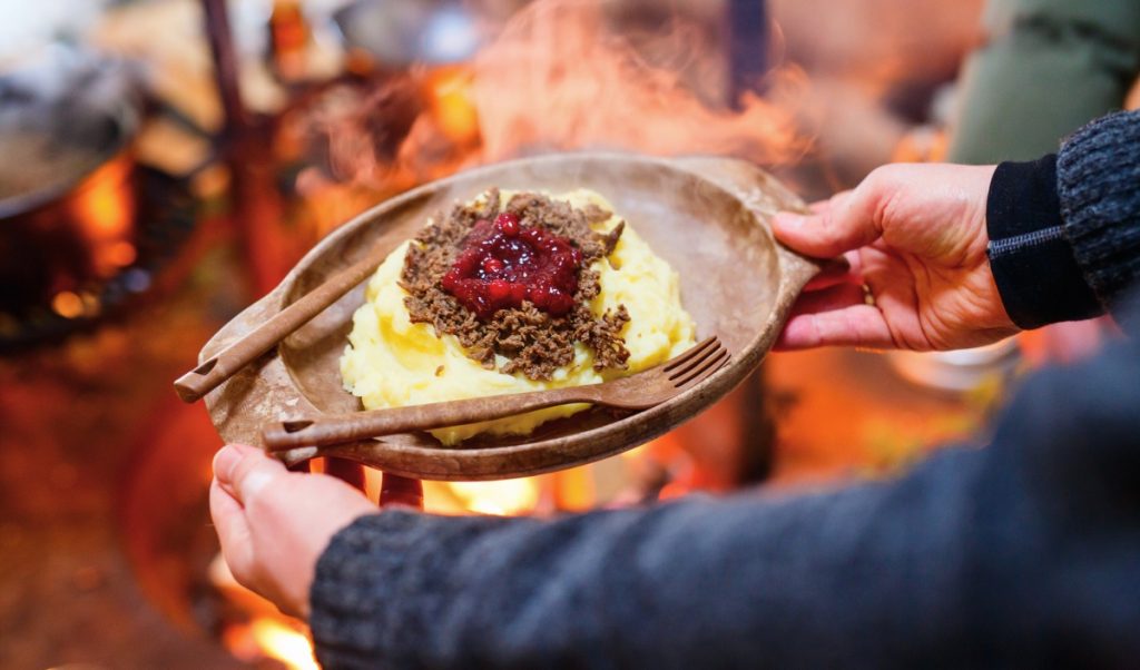 A traditional Finnish meal of mashed potatoes topped with minced meat and lingonberries is served on a wooden plate. Steam rises from the food as someone holds it near an open fire.