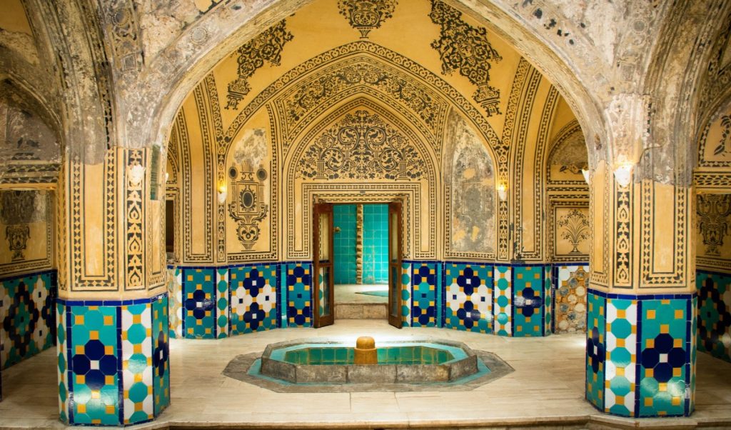 Ornate hammam interior with turquoise, blue, yellow, and white tilework around a small central fountain. Arched walls and columns are covered in faded black and gold decorative patterns.