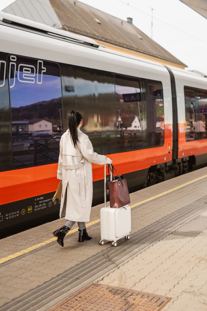 Woman in a cream coat rolling a white suitcase along a train platform beside an orange and black train. She carries a tote bag and looks toward the train doors while traveling by rail.