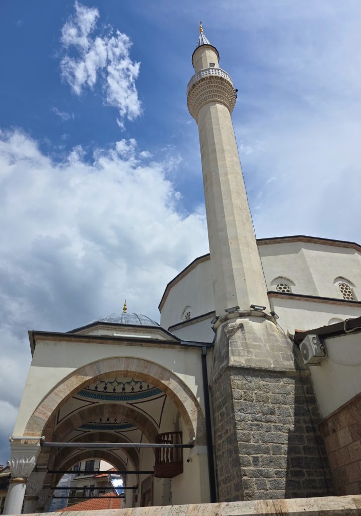 A tall white minaret rises above an arched mosque courtyard in Ohrid under a partly cloudy sky.