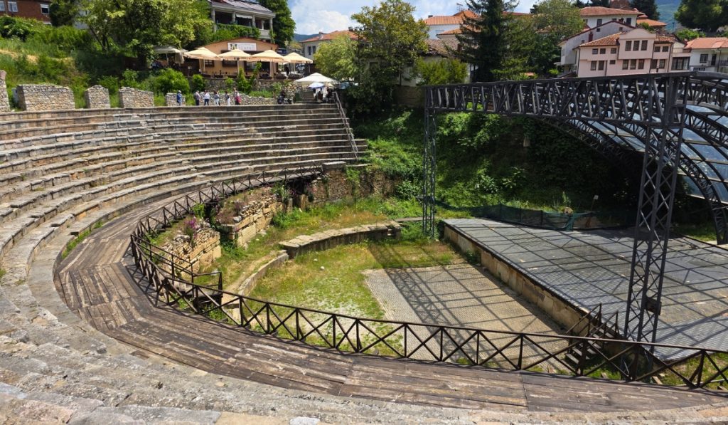 Stone seating curves around the ancient theatre in Ohrid with a black stage structure and town buildings above it.