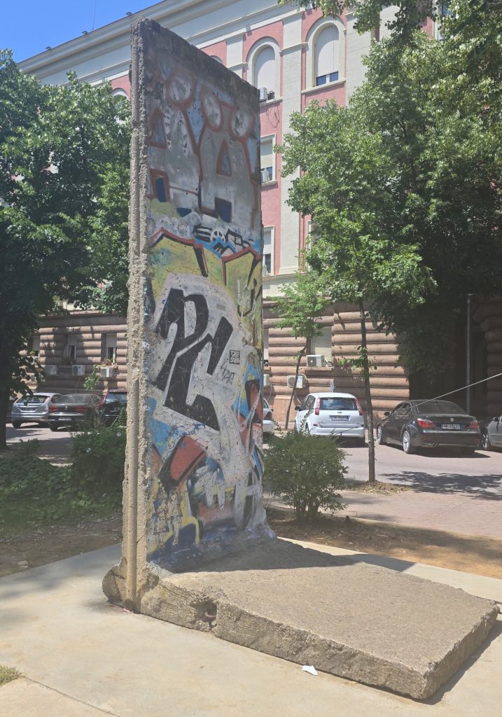 Graffiti covered section of the Berlin Wall standing on a concrete base in Tirana. Trees. parked cars. and a red and white building sit behind the wall fragment.