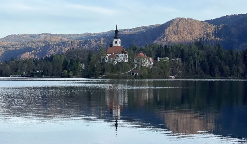 Bled Island church reflects in the calm water of Lake Bled during the day. Forested hills and mountains surround the lake behind the island.