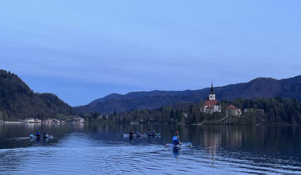 Small groups kayak across Lake Bled toward Bled Island on a calm evening. The island church sits near the shore with wooded hills and soft blue sky behind it.