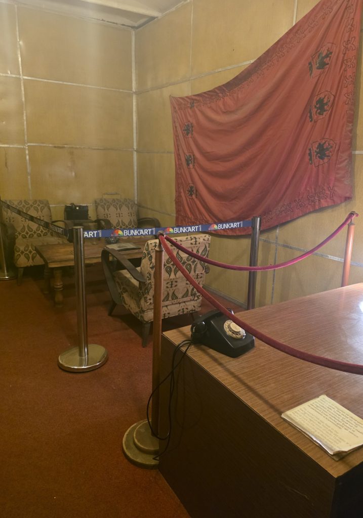 Preserved room behind ropes at Bunk Art with patterned armchairs. a wooden desk. a black rotary phone. and a red Albanian flag hanging on the wall. The barrier tape reads "BUNKART 1."