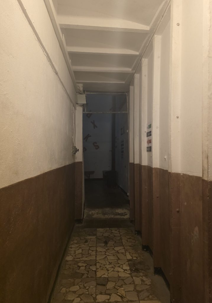 Narrow bunker corridor with brown and cream walls leading toward a dark open doorway. The worn stone floor and low ceiling make the passage feel enclosed.