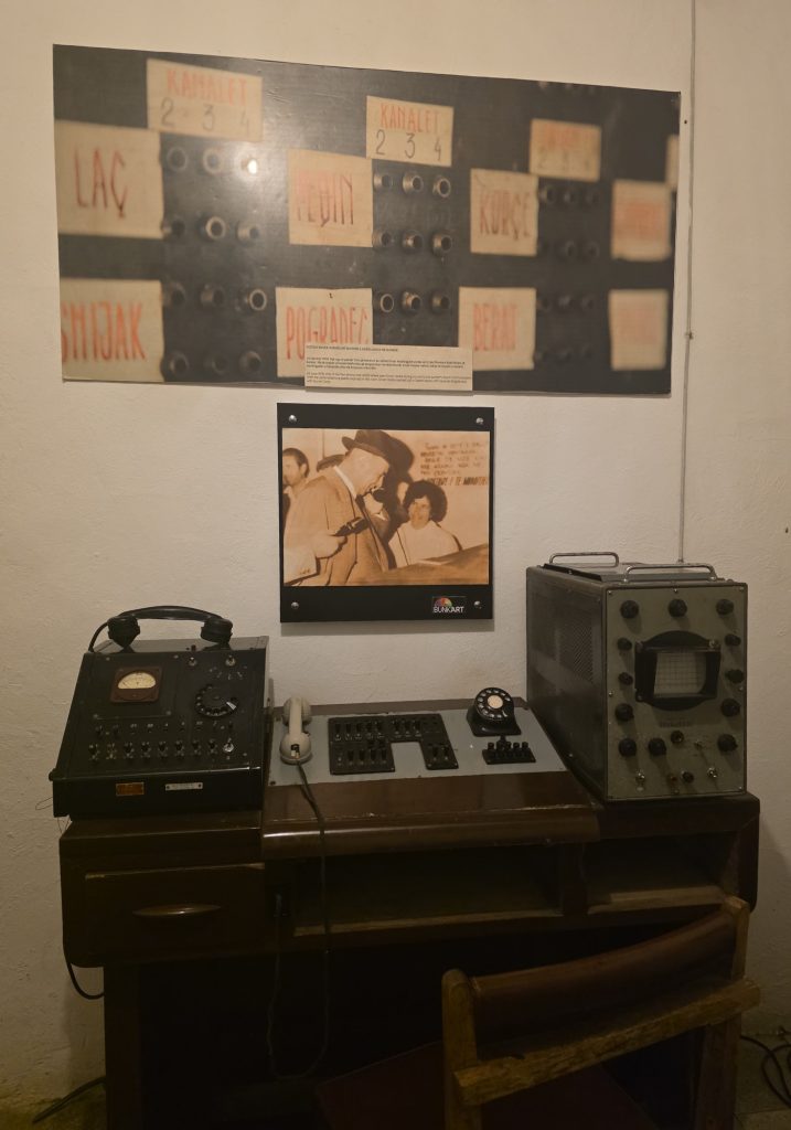 Museum display of vintage communications equipment on a wooden desk with wall photos above it. The upper photo includes labels reading "LAC." "KANALI 2 3." "TË GJIN." "KANALI 2 3 4." "KORÇË." "POGRADEC." and "REZA."