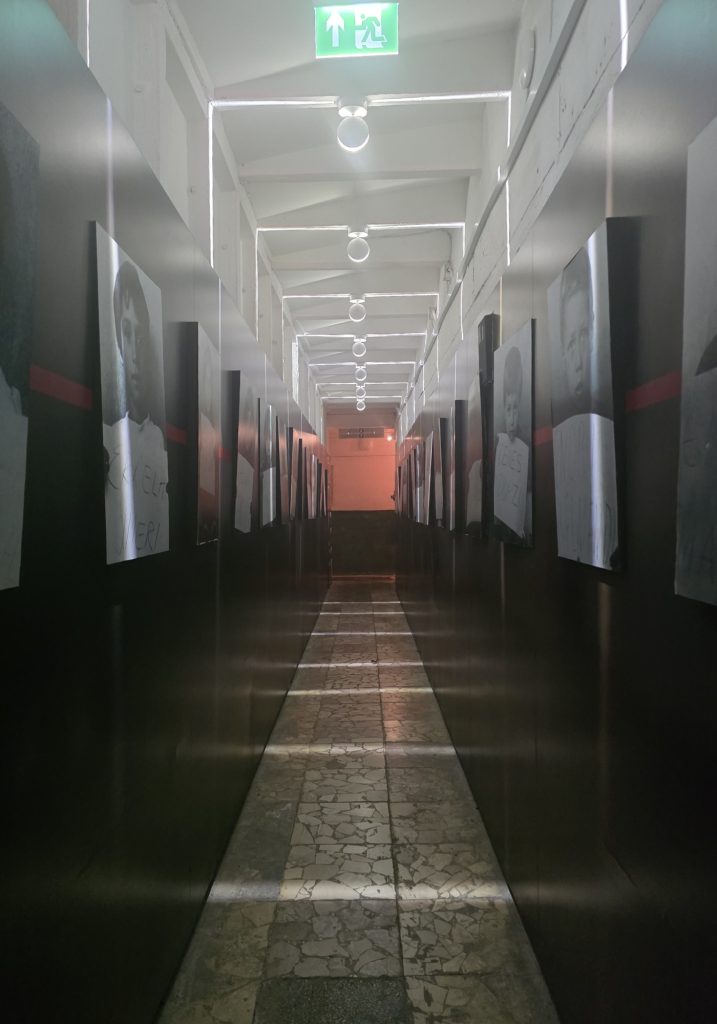 Long museum corridor lined with black and white portraits on both walls. A green exit sign glows overhead above the narrow tiled walkway.