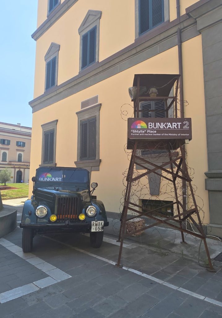 Exterior of Bunk Art 2 with a black vintage vehicle parked beside a barbed wire watchtower. The sign reads "BUNK ART 2." "Shtylla Place." and "Former anti nuclear bunker of the Ministry of Interior."