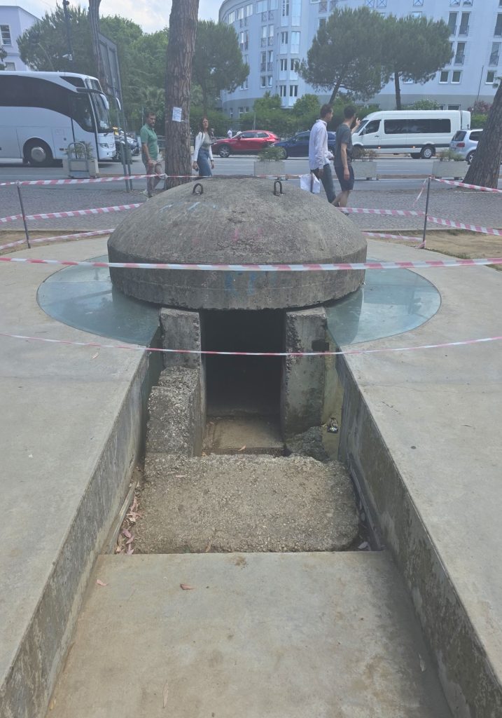 Small concrete bunker entrance with a domed roof blocked off by red and white tape. Pedestrians. buses. and city traffic pass behind it on a tree lined street.