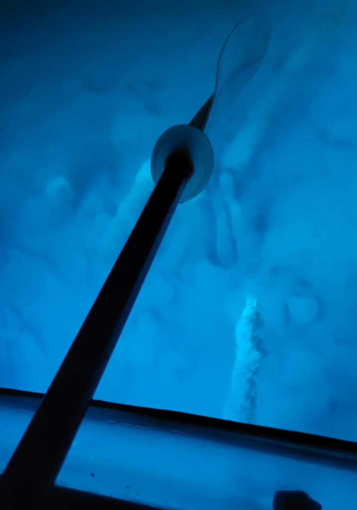 Kayak paddle rests over glowing blue water seen through the clear bottom of a kayak. Pale rocks and shadows are visible beneath the lake surface.