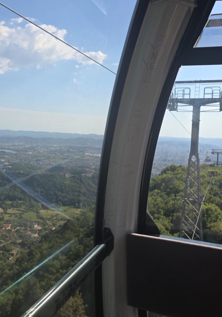 View from inside a cable car with a city and green hills visible through the window. The window frame has scratched writing including "2022." and a heart with "N E."