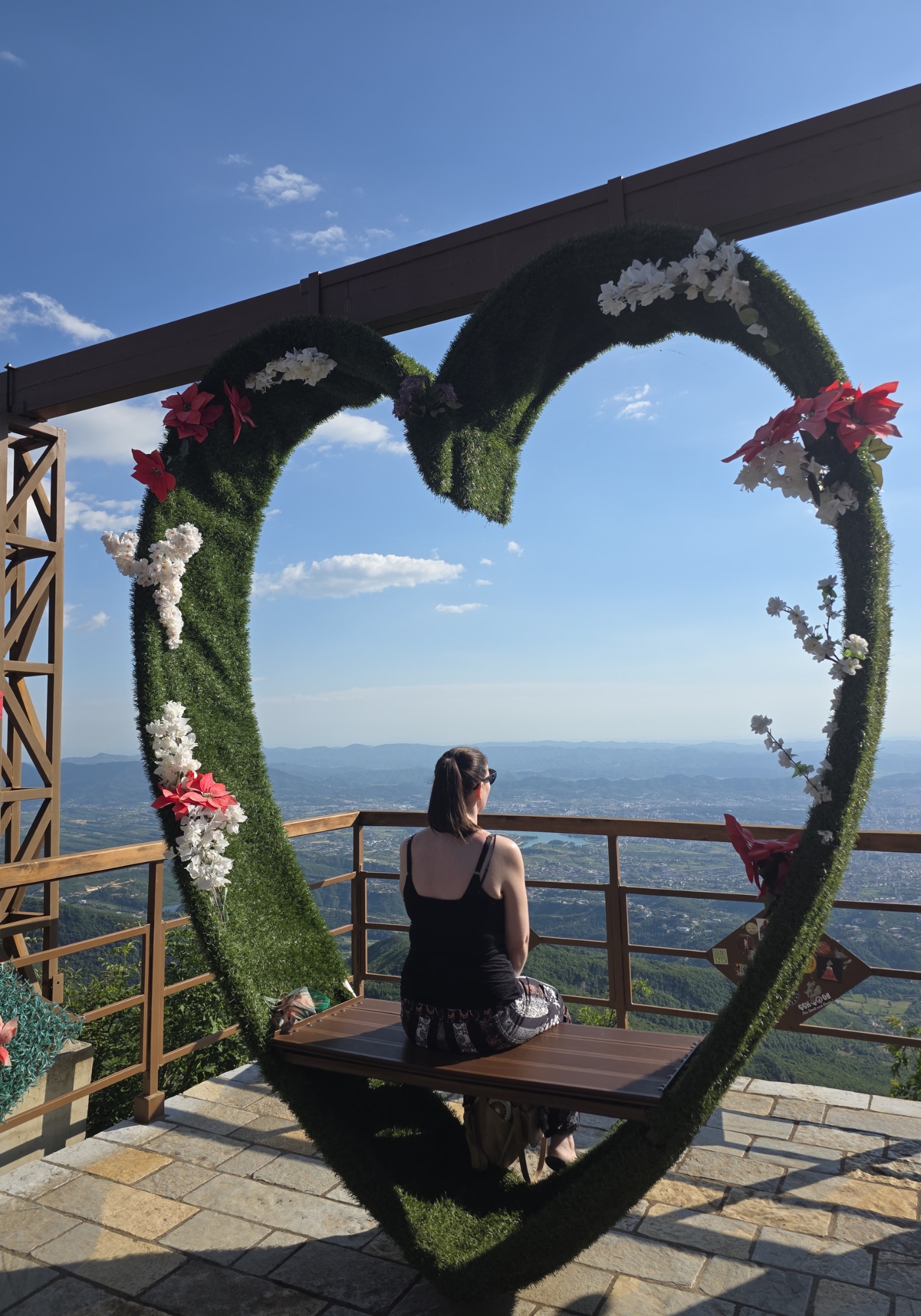 Sitting on a bench inside a large heart shaped greenery frame decorated with red and white flowers. The viewpoint looks out over distant mountains and a valley under a bright blue sky.