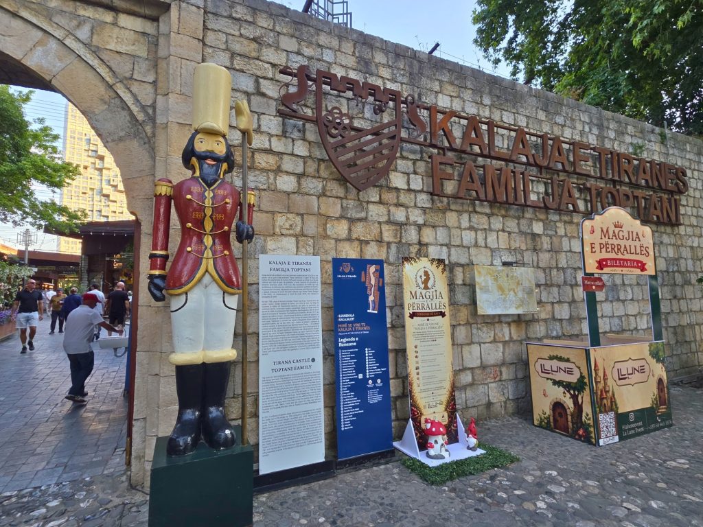 Stone entrance wall for Tirana Castle with a toy soldier statue and tourist signs in front. Large lettering reads "Kalaja e Tiranës Familja Toptani." with signs for "Tirana Castle Toptani Family." and "Magjia e Përrallës Biletaria."