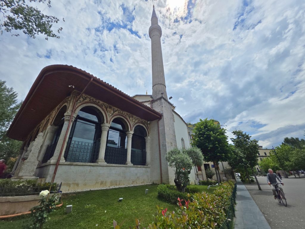Historic mosque with arched windows and painted trim beside a tall minaret. A cyclist rides past the garden and walkway under a cloudy blue sky.