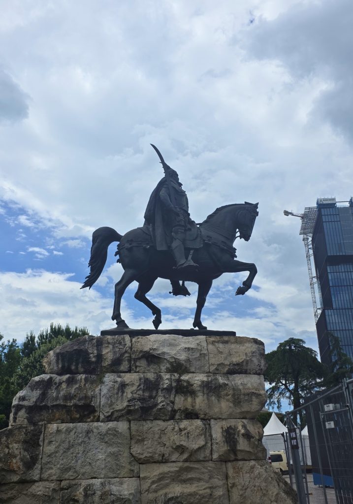 Dark equestrian statue of a warrior holding a curved sword on a stone pedestal. Modern buildings and construction fencing surround the monument under a cloudy sky.