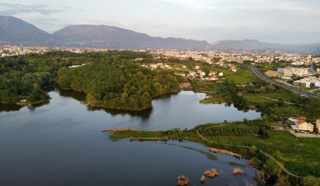 Aerial view of a lake bordered by dense green trees with Tirana spread out beyond it. Mountains rise in the distance under a soft evening sky.