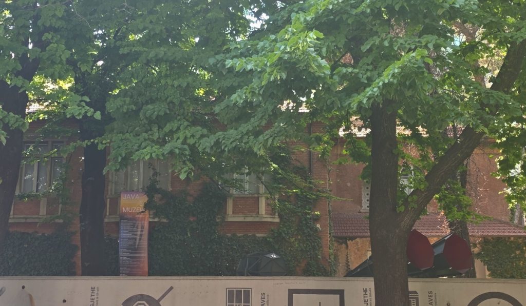 Leafy trees shade an old brick building partly covered in vines. A sign in front begins with "JAVA" and "MUZE." while construction panels line the sidewalk.