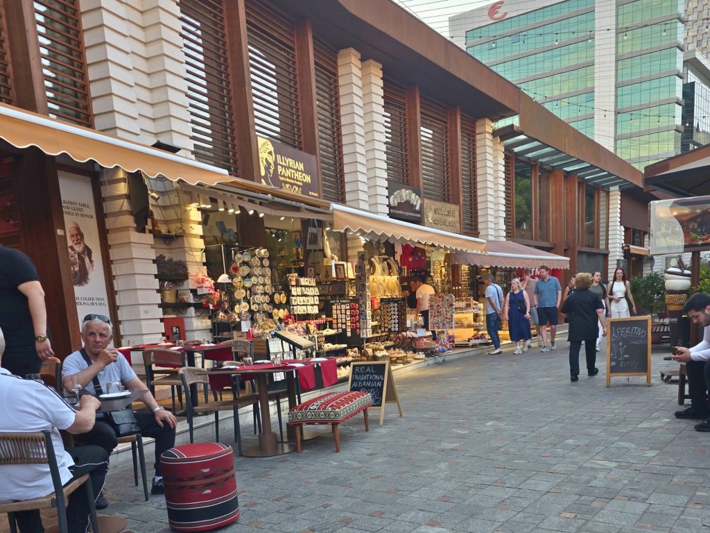 Pedestrian shopping lane with souvenir stalls. cafe tables. and people walking past restaurants near Tirana Castle. Signs read "Illyrian Pantheon." "Real traditional Albanian food." and "Aperitivo."