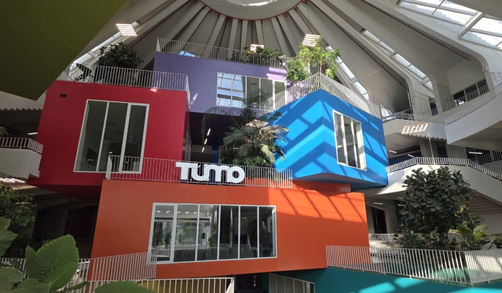 Colorful stacked rooms inside the Pyramid of Tirana with a white "Tumo" sign on the orange level. Skylights. plants. railings. and staircases fill the bright atrium.